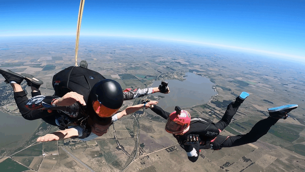Salto tándem en caída libre con camarógrafo, instructor y pasajero tomados de la mano sobre el cielo de Chascomús